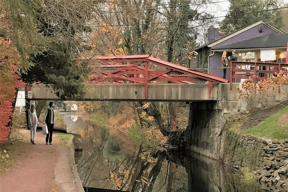 A scenic canal path with a red bridge, surrounded by autumn foliage and two people walking.