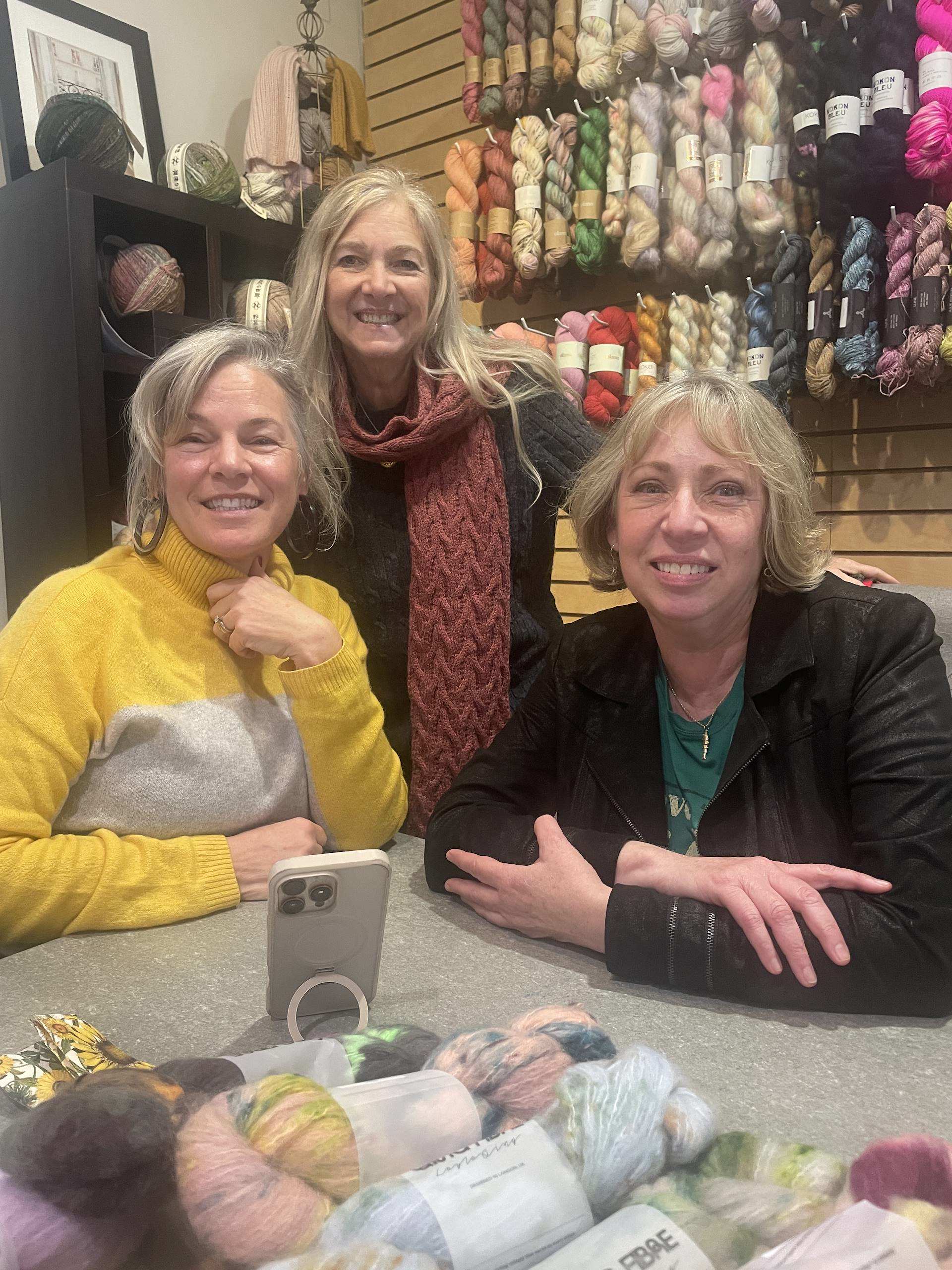 Three women are smiling together in a yarn shop, with colorful yarn displayed in the background.