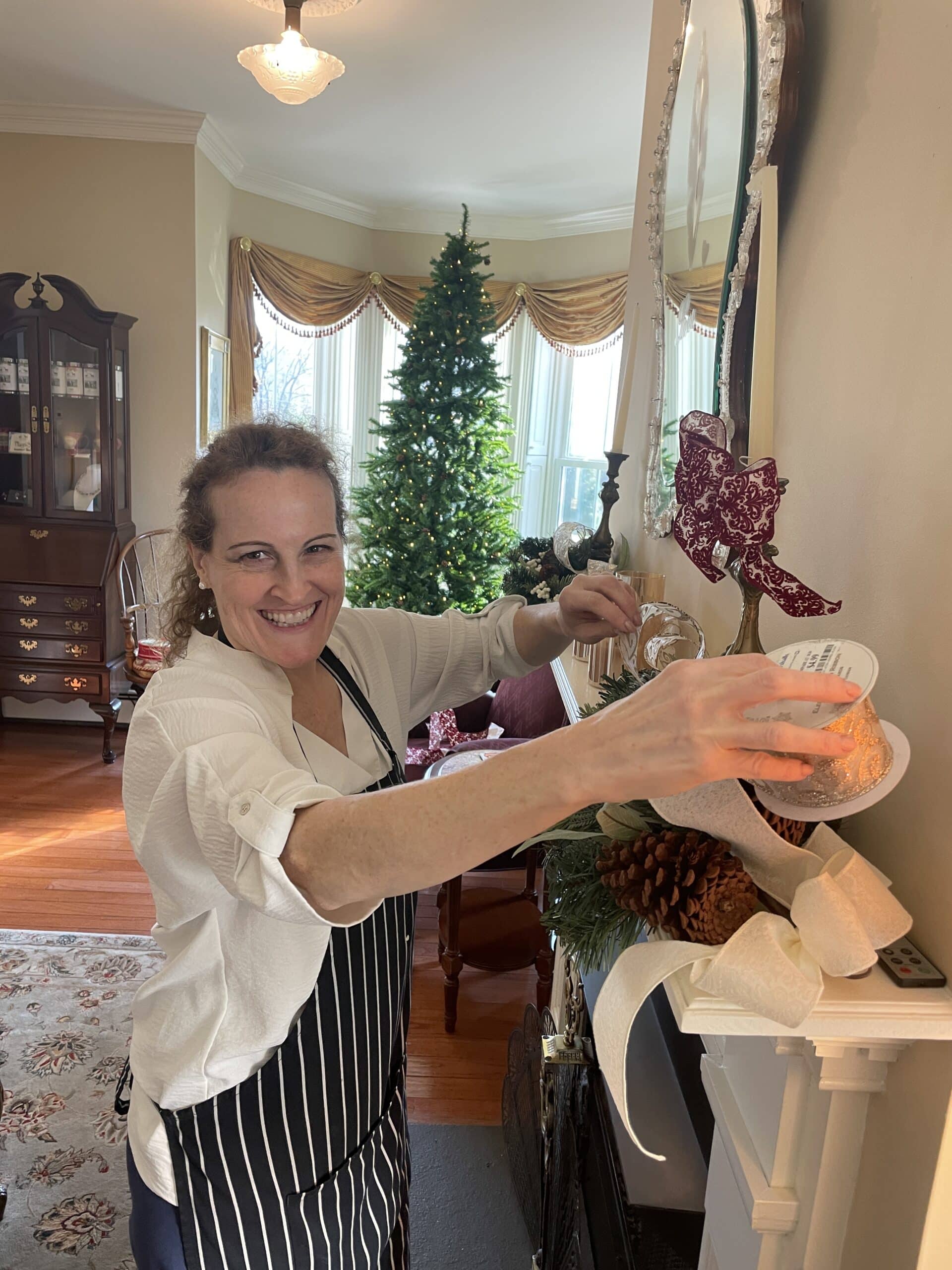A woman in a striped apron decorates a mantel with festive items while smiling in a beautifully arranged room.