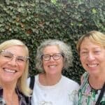 Three smiling women take a selfie in front of a green, leafy background.