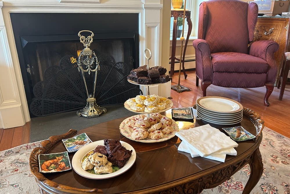A table displays an assortment of baked goods alongside plates and napkins in a cozy room with a fireplace.