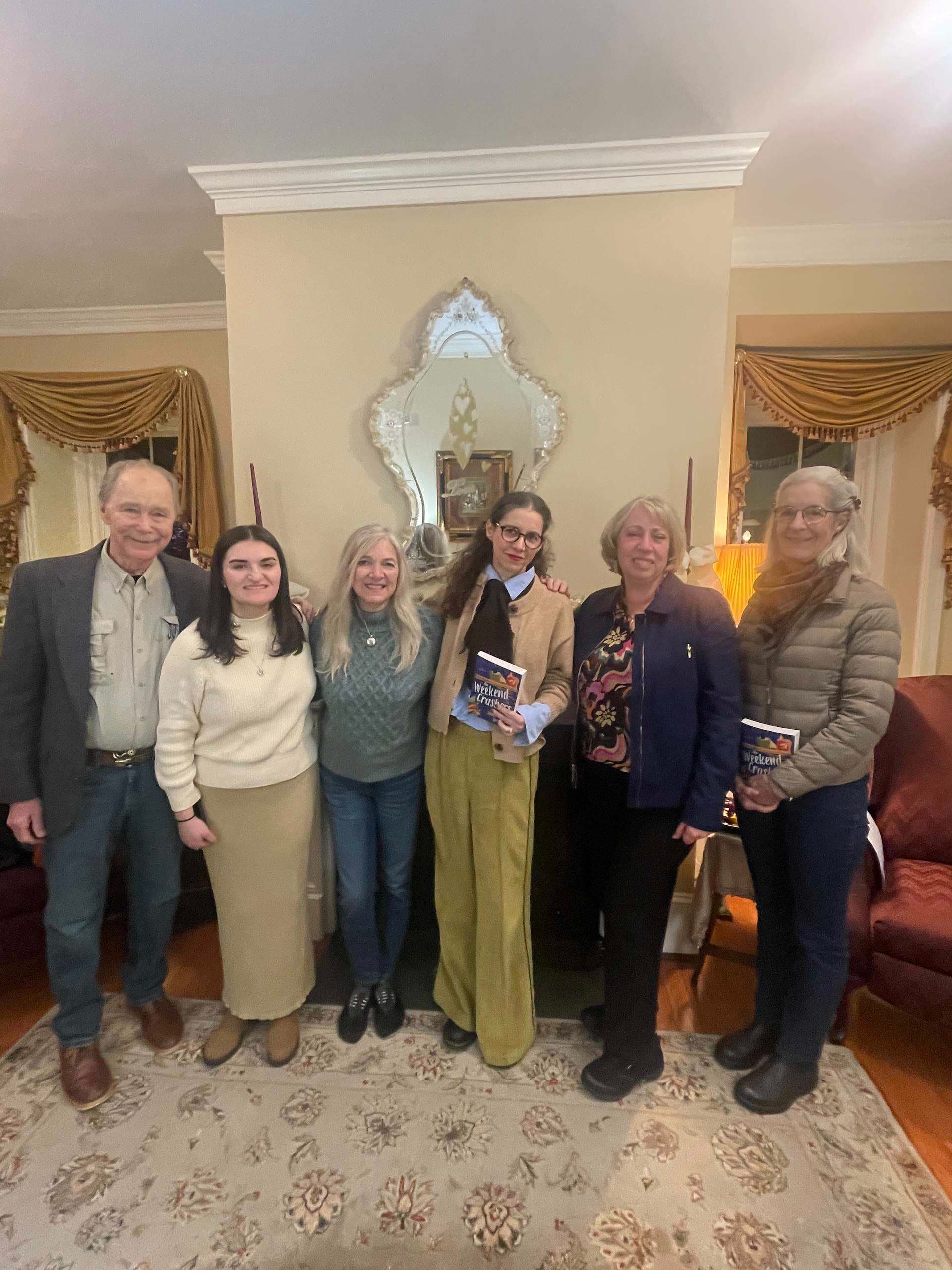 A group of six people, standing together in a cozy living room, smiling and holding a book.