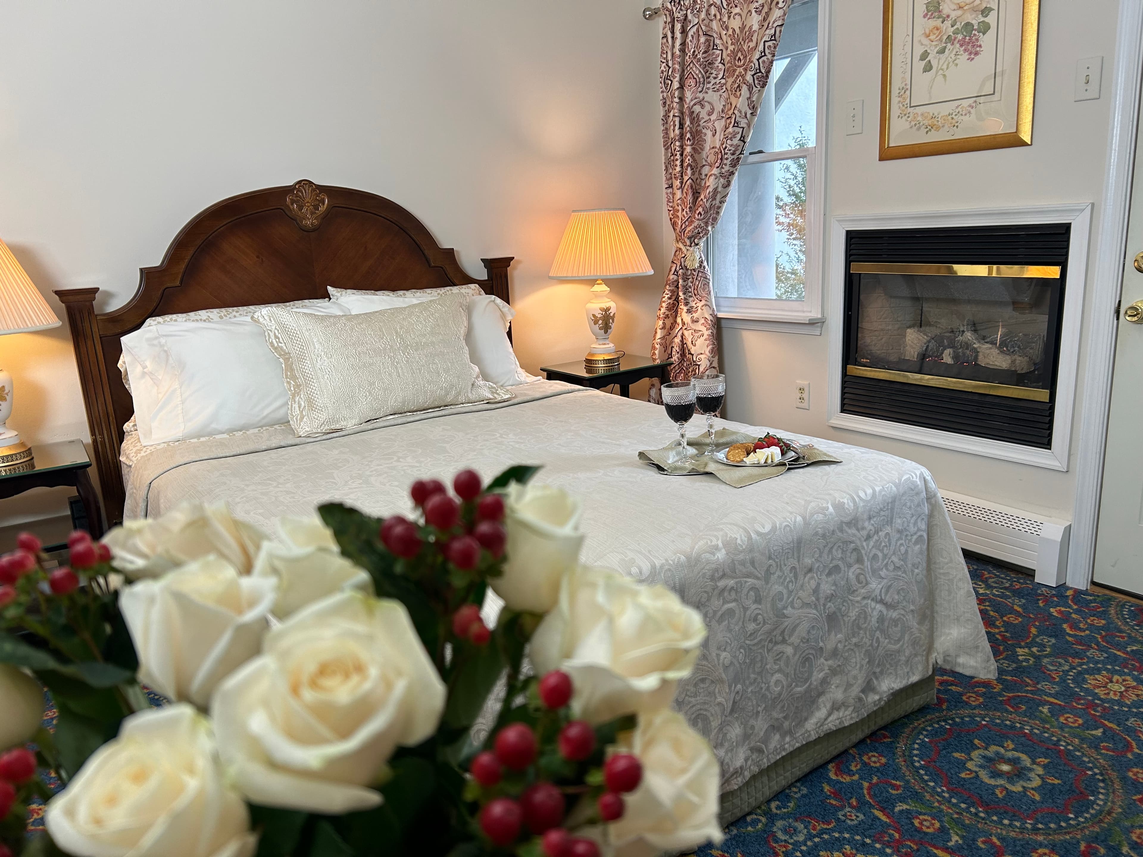 A cozy bedroom featuring a neatly made bed, decorative pillows, a fireplace, and a bouquet of white roses in the foreground.