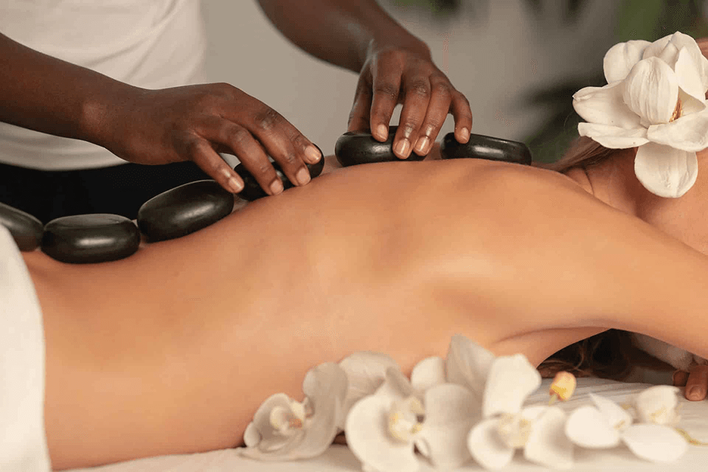 A person receives a hot stone massage surrounded by white orchids.