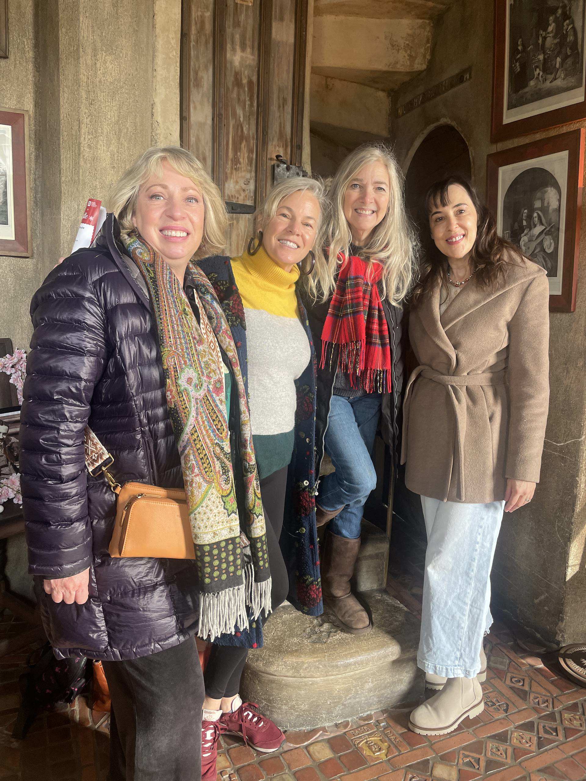 Four women stand together in a cozy, rustic interior, smiling and dressed in winter attire.