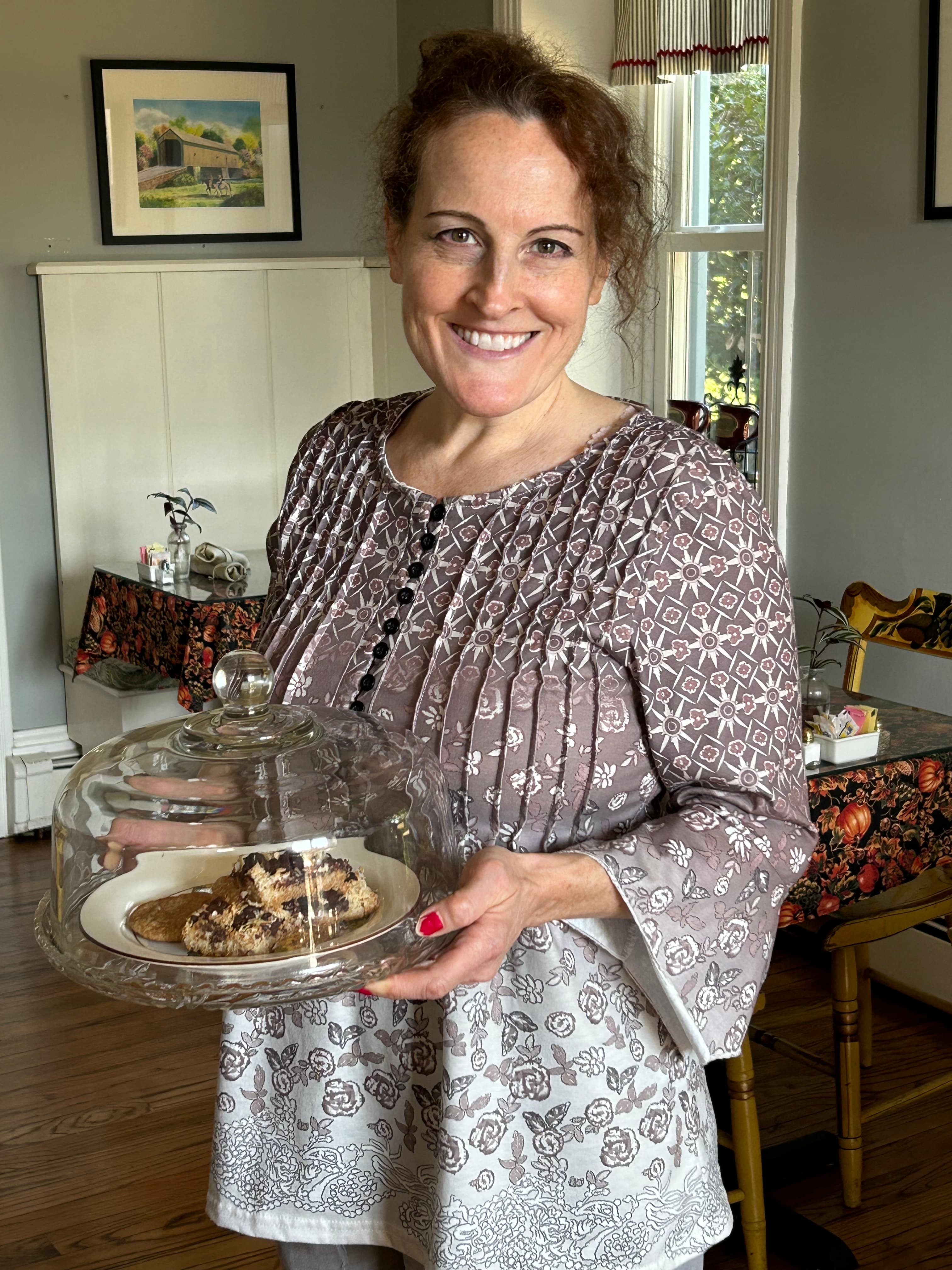 A smiling woman holds a covered dessert on a plate in a cozy dining room.