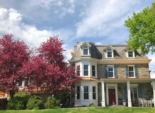 A charming stone house with a red door, flanked by blooming pink trees and green lawn under a partly cloudy sky.