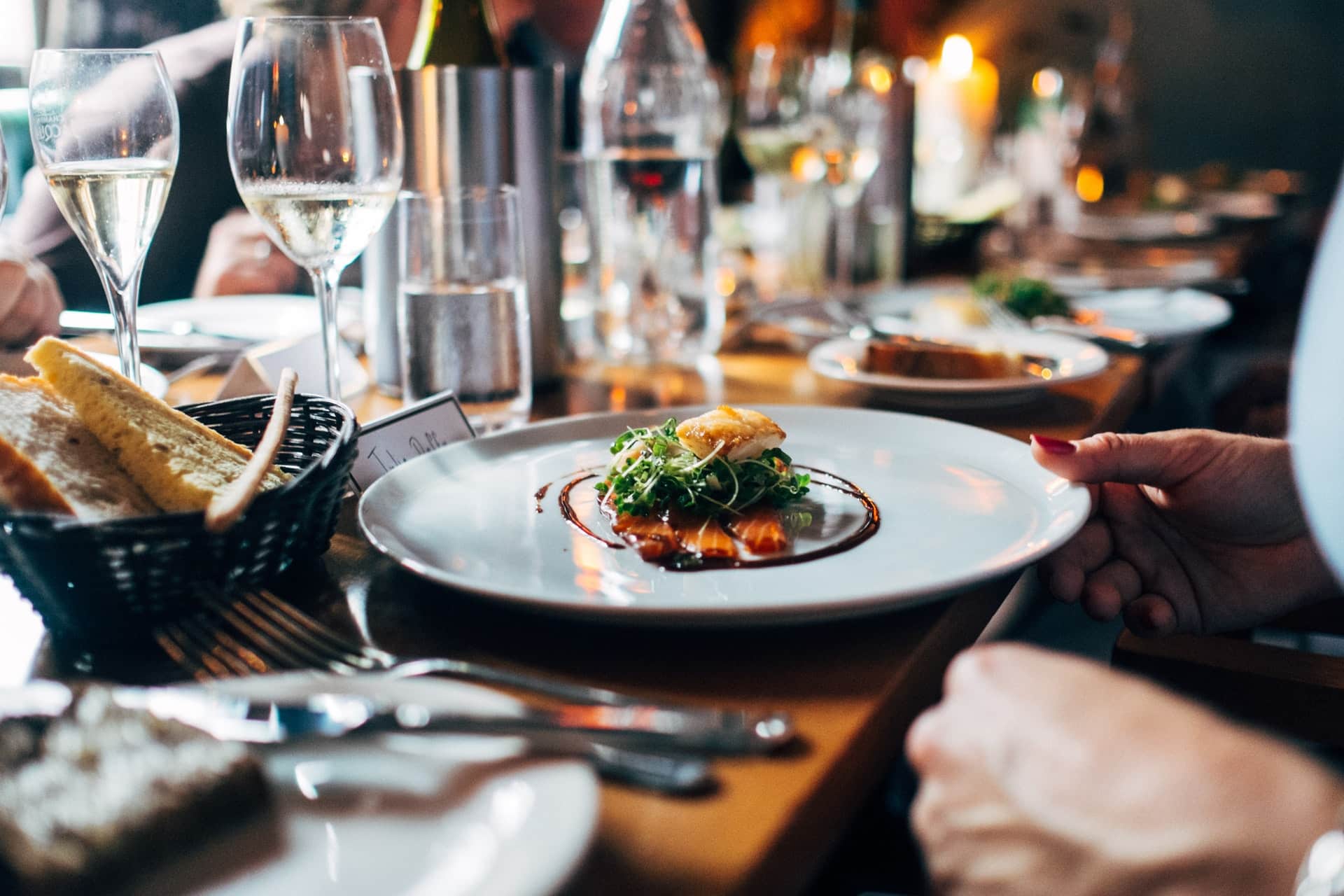 A elegantly plated dish with greens and sauce is being served at a dining table surrounded by glasses and a basket of bread.