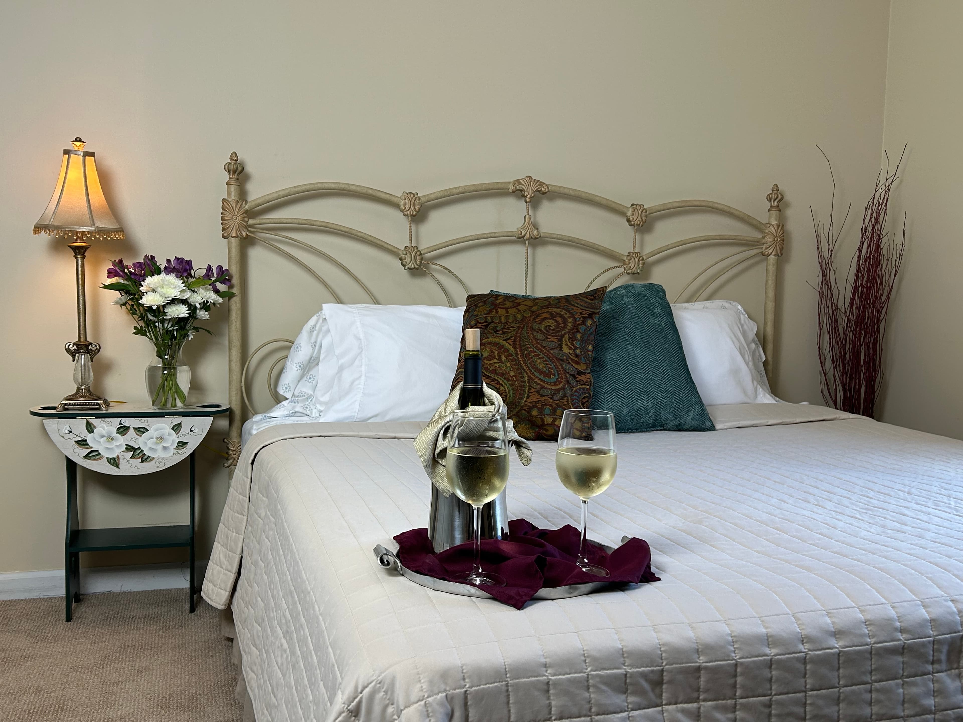 View of king bed, side table & lamp with wine & wine glasses on bed