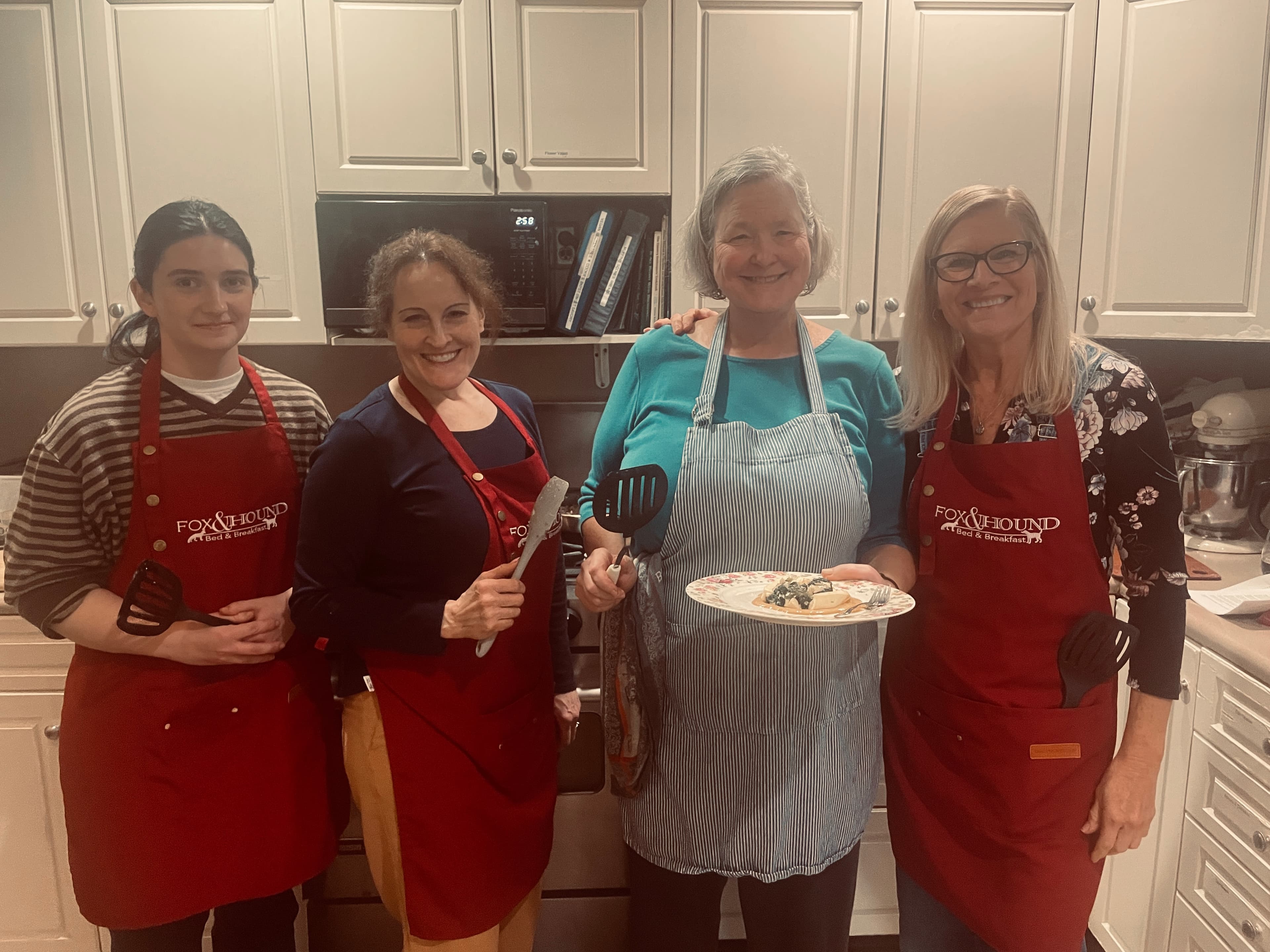 Four women wearing aprons stand together in a kitchen, smiling and holding cooking tools and a plate of food.