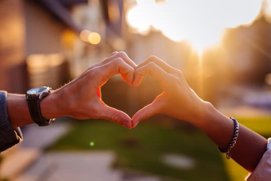 Two hands forming a heart shape against a sunset backdrop.