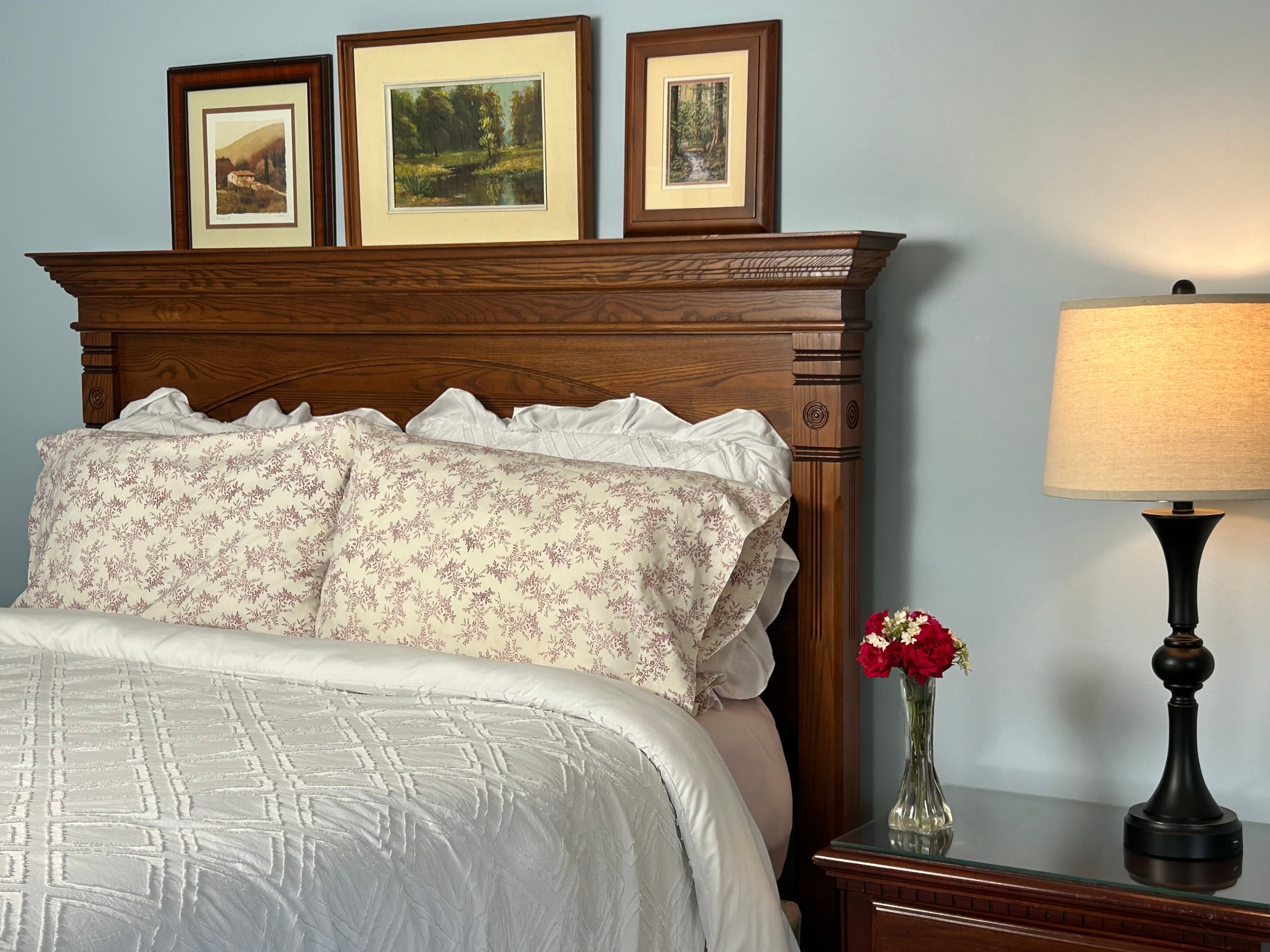 View of queen bed with wood headboard, side table, lamp with roses