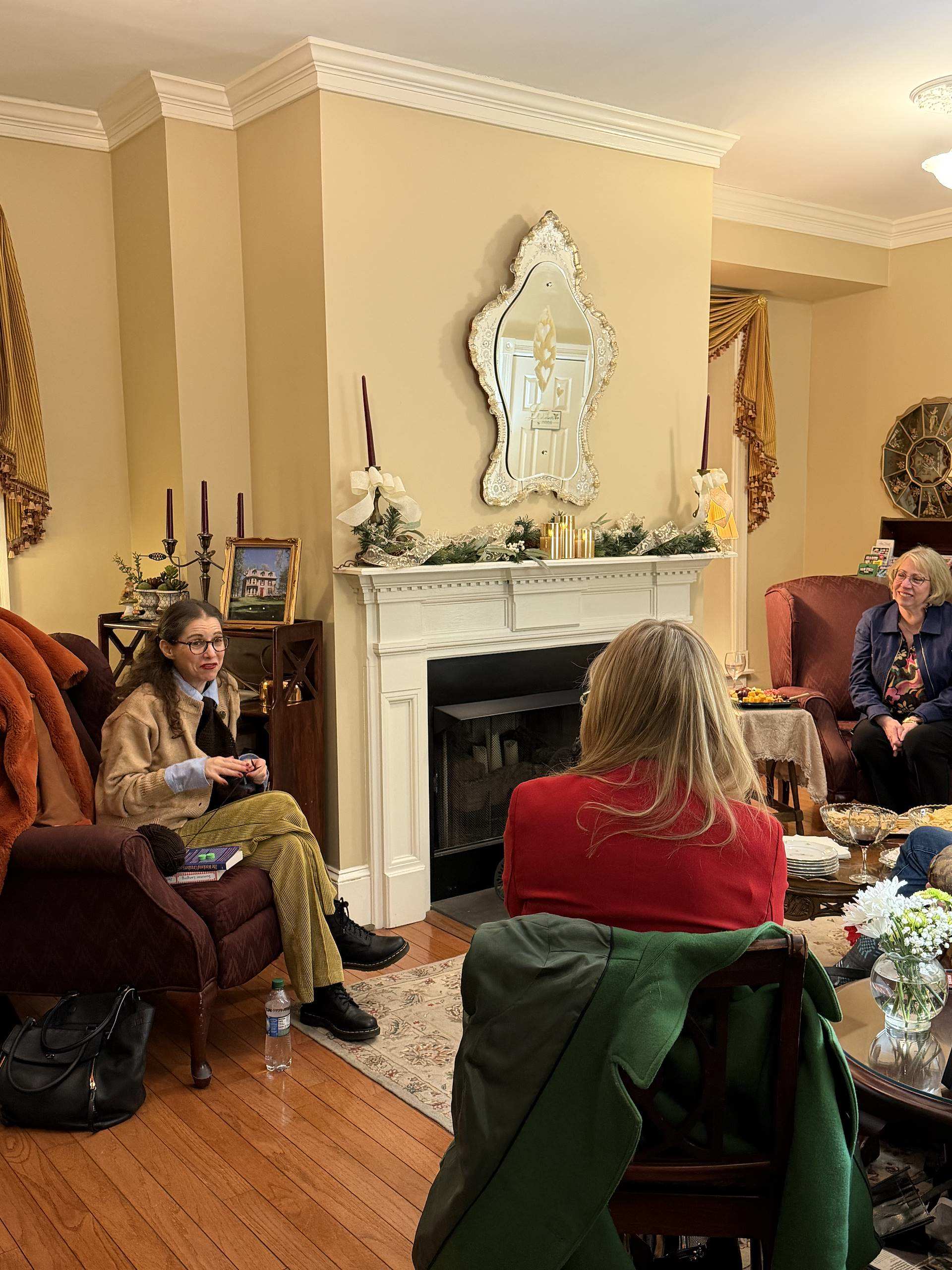 A woman in a brown coat speaks in a cozy living room while others listen attentively.