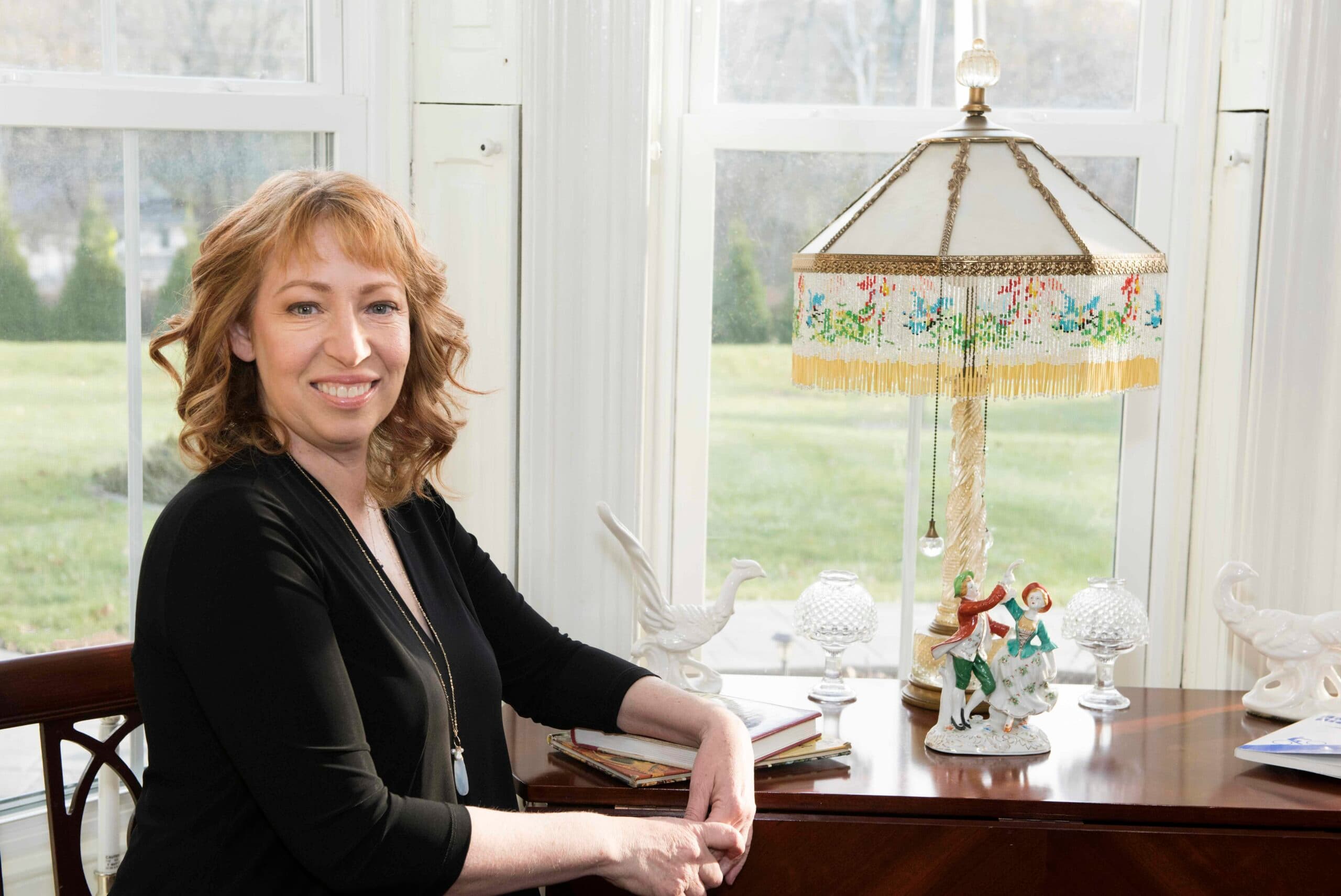 A woman with wavy hair smiles while seated by a lamp on a wooden desk, surrounded by decorative figurines and sunlight filtering through large windows.