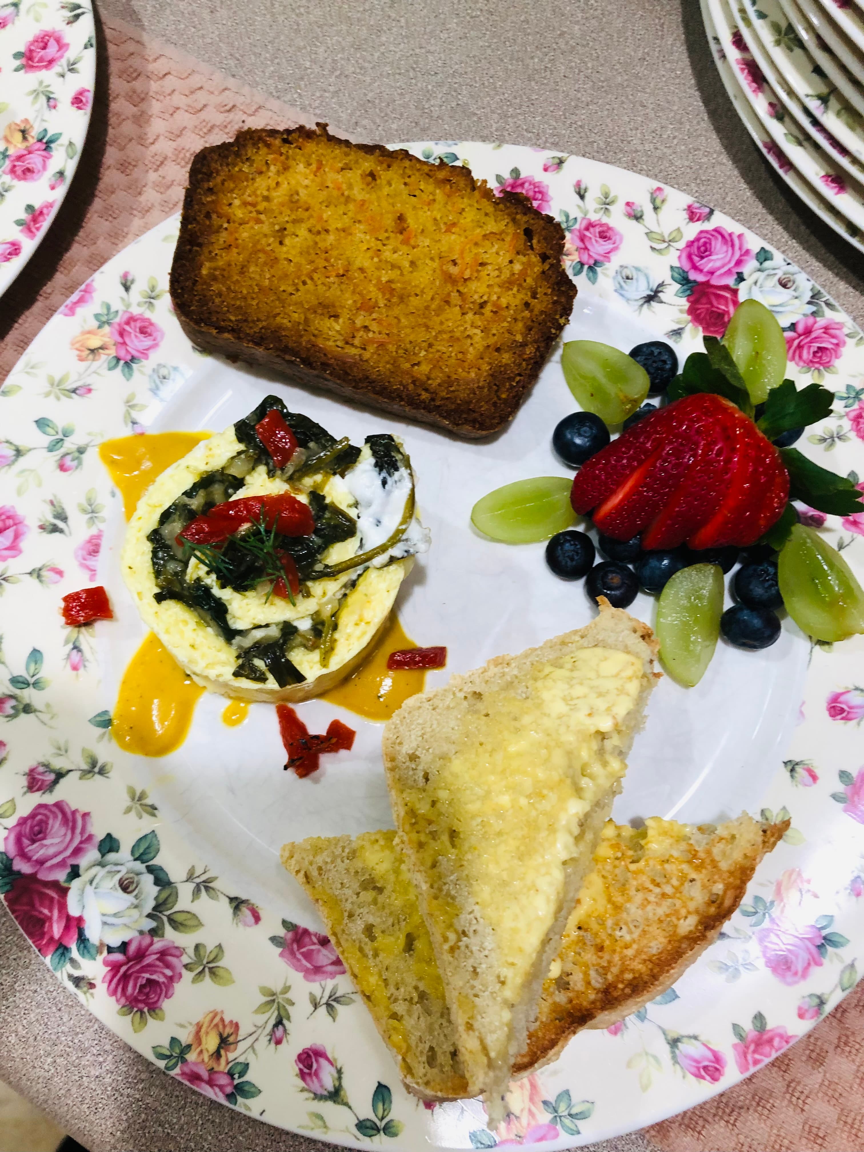 A floral plate featuring a slice of cornbread, a vegetable egg dish, two pieces of toast, and a variety of fresh fruit.